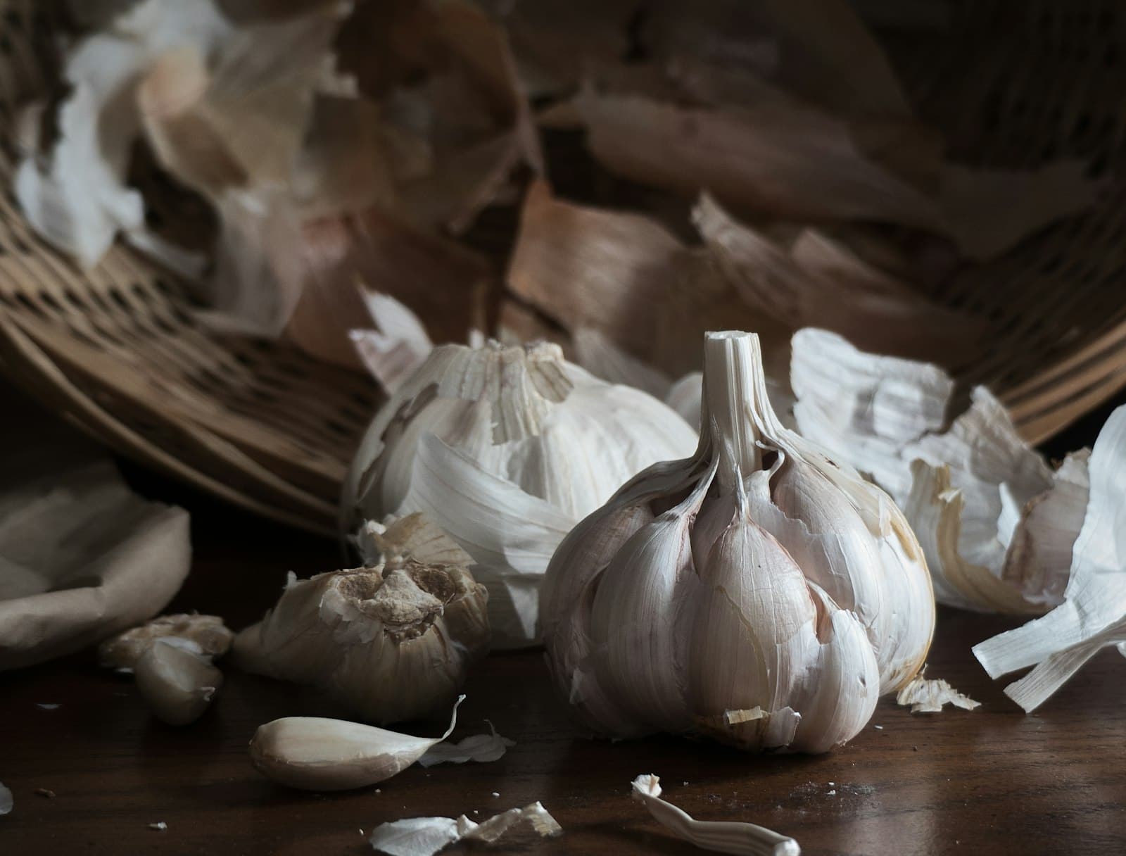 Cured garlic bulbs and loose cloves in a woven basket on a dark wooden table.